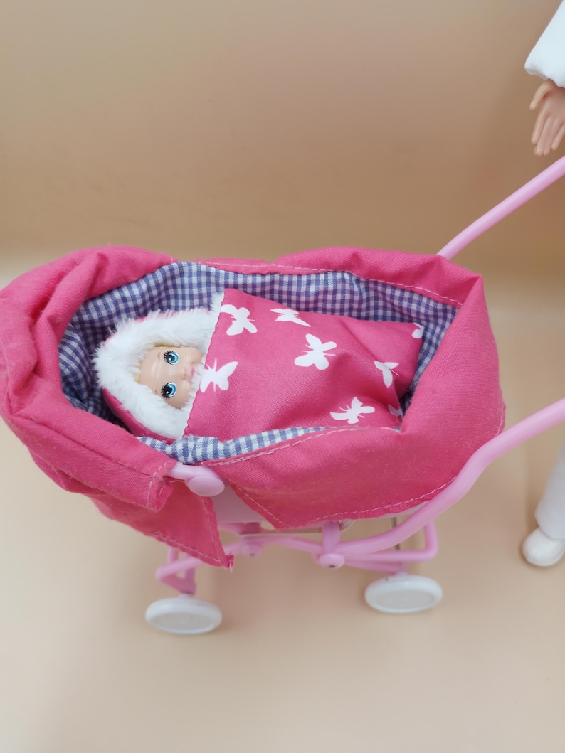 May include: A pink doll stroller with a baby doll inside. The baby is wrapped in a pink blanket with white butterfly designs. The stroller has white wheels and a pink handle.