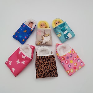 May include: Six doll sleeping bags in assorted designs and colours. Patterns include stars, butterflies, a unicorn, a snowman, leopard print, and polka dots. Each bag features a white, fluffy interior.