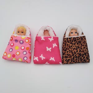 May include: Three doll sleeping bags in pink, leopard print, and a pink pattern with white butterflies. Each bag has a white fluffy trim and a doll's face peeking out. A fun toy for children.