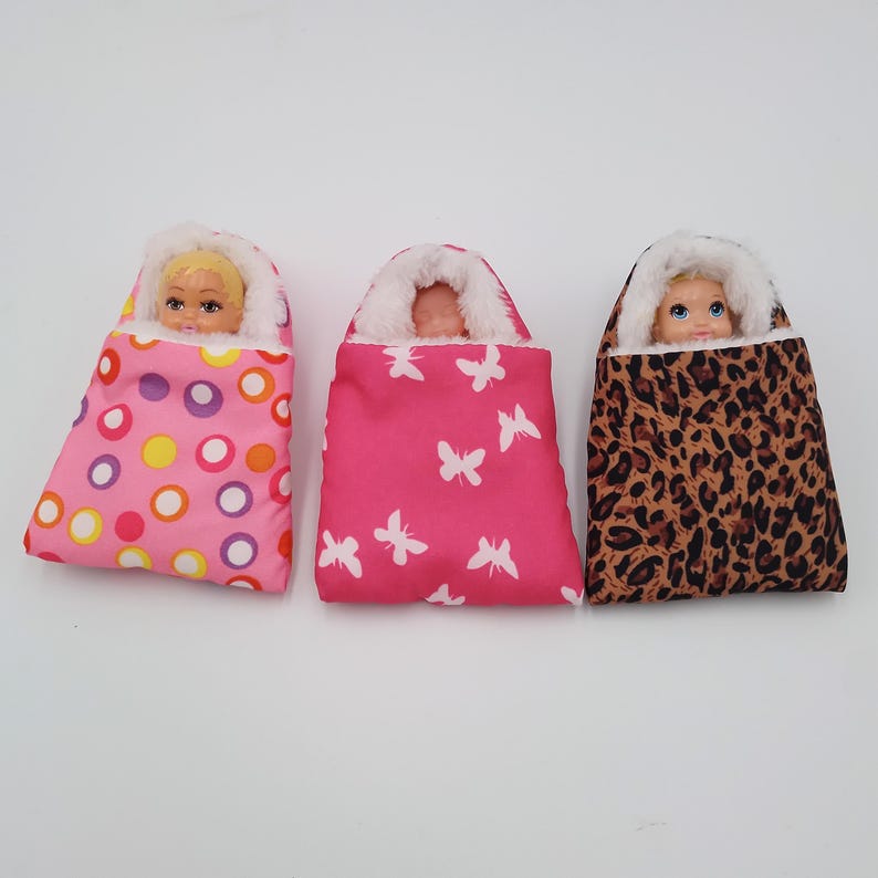 May include: Three doll sleeping bags in pink, leopard print, and a pink pattern with white butterflies. Each bag has a white fluffy trim and a doll's face peeking out. A fun toy for children.