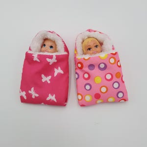 May include: Two doll-sized baby sleeping bags. One is hot pink with white butterfly shapes, and the other is pink with a pattern of colorful circles. Each bag has a white fluffy trim and a doll's head peeking out.