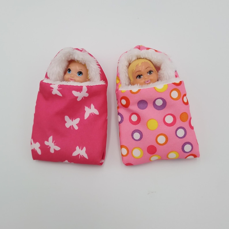 May include: Two doll-sized baby sleeping bags. One is hot pink with white butterfly shapes, and the other is pink with a pattern of colorful circles. Each bag has a white fluffy trim and a doll's head peeking out.