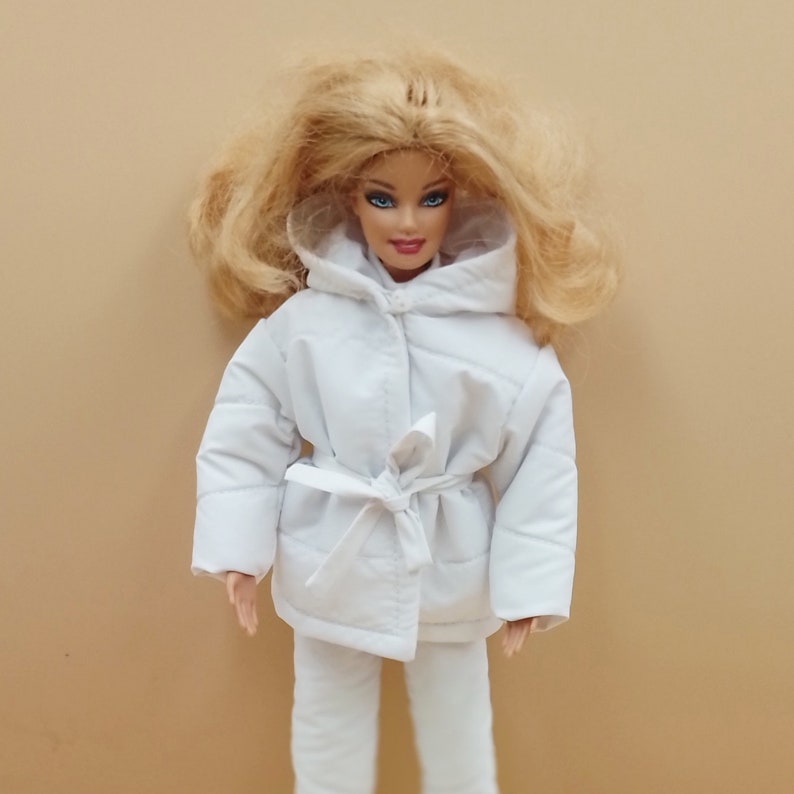 K&ouml;nnte beinhalten: Eine wei&szlig;e Kapuzenjacke mit einem Bindeg&uuml;rtel. Die Jacke ist aus gestepptem Stoff und hat einen Rei&szlig;verschluss. Die Puppe tr&auml;gt ein wei&szlig;es Outfit.
