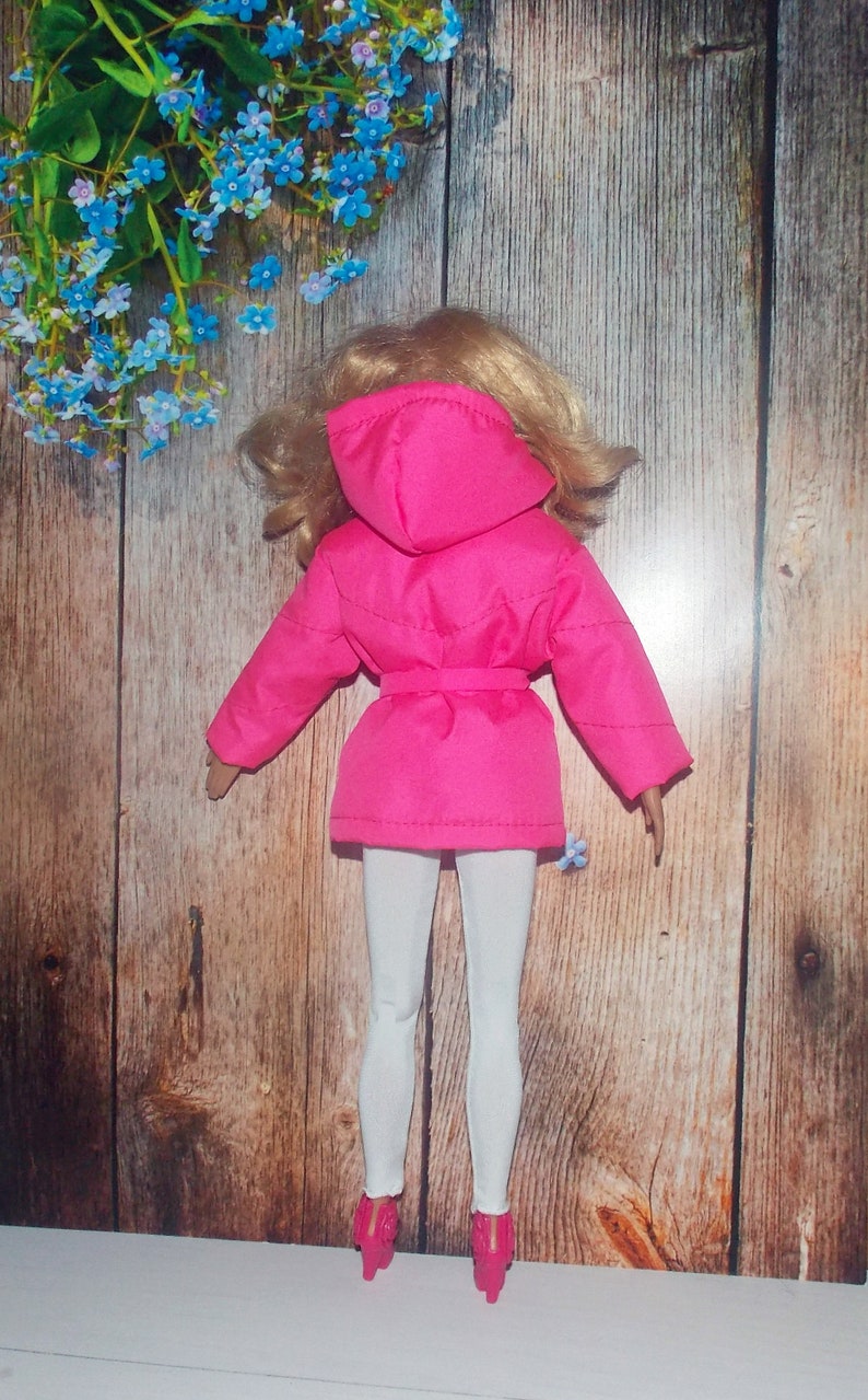 K&ouml;nnte beinhalten: Eine rosa Kapuzenjacke mit G&uuml;rtel und wei&szlig;er Hose. Die Jacke hat ein gestepptes Design und wird von einer Puppe getragen.