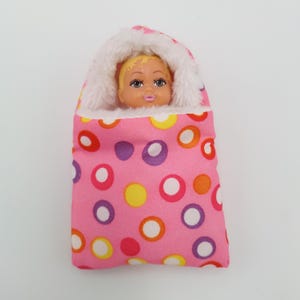 May include: A doll with blonde hair peeks out of a pink baby bunting. The bunting is decorated with colorful polka dots and lined with white faux fur. The baby doll has painted facial features.