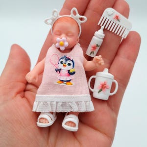 Può includere: Una bambola neonato in miniatura che indossa un vestito rosa con grafica di pinguino, una fascia bianca e sandali bianchi. Il set include un biberon bianco, un bicchiere bianco con fiore rosa e un pettine bianco con piccolo fiore rosa.