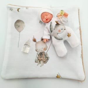 May include: A small baby doll wearing a white outfit and hat, lying on a white blanket with a whimsical print. The blanket features illustrations of a balloon, a bunny on a swing, and stars. A small bottle is also present.