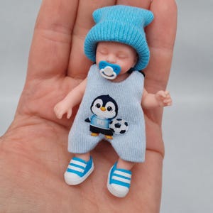 以下が含まれることがあります： 水色のニット帽と、サッカーボールを持ったペンギンのグラフィックが描かれた水色のニットロンパースを着たミニチュアの赤ちゃん人形。人形は水色のおしゃぶりをしており、小さな白と青のスニーカーを履いています。