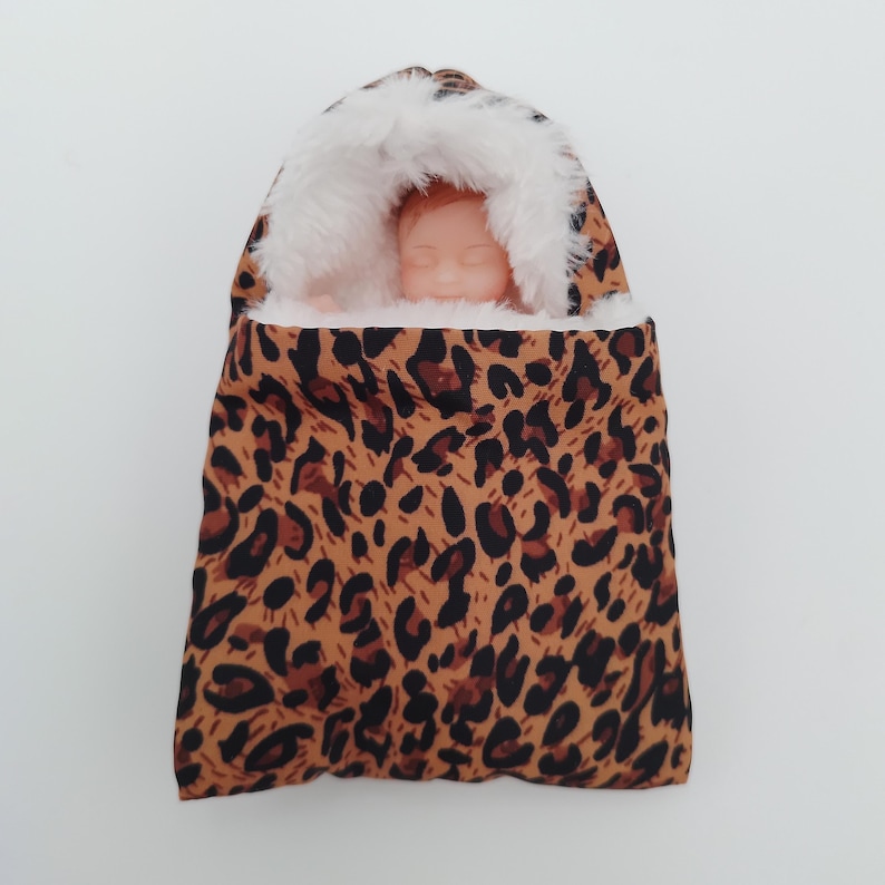 May include: A doll swaddled in a leopard print baby bunting. The bunting has a brown and black spotted pattern with a white faux fur lining around the hood. The doll has a peach skin tone and is nestled inside the bunting.