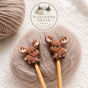 May include: Two light brown wooden knitting needles, each topped with a small, flat, brown moose-shaped embellishment featuring cream-colored antlers, black eyes, and black hooves, positioned in front of a ball of light brown yarn.