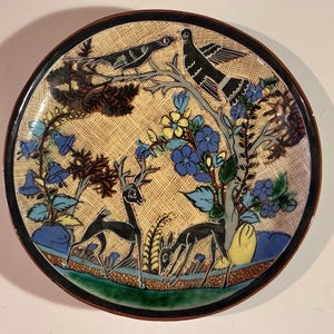 May include: A round ceramic plate featuring a hand-painted scene of two black deer standing in a grassy field beneath a tree with blue and yellow flowers. Two black birds are perched on the branches above. The background has a textured tan crosshatch pattern.