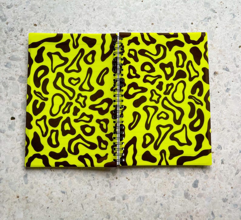 May include: A yellow notebook with a black and yellow abstract pattern on the cover. The notebook is closed and has a spiral binding.
