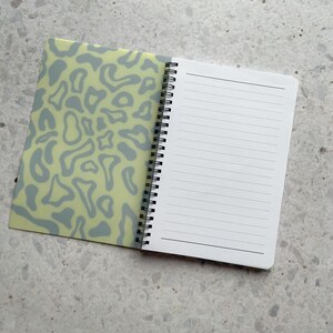 May include: A spiral bound notebook with a light green cover featuring a gray abstract pattern. The notebook has white lined pages.