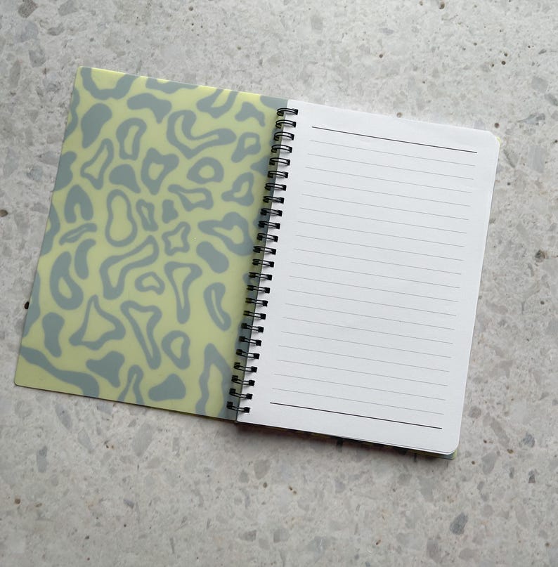 May include: A spiral bound notebook with a light green cover featuring a gray abstract pattern. The notebook has white lined pages.