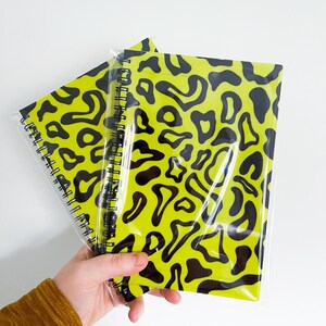 May include: Two spiral bound notebooks with black and yellow abstract patterns. The notebooks are wrapped in clear plastic.