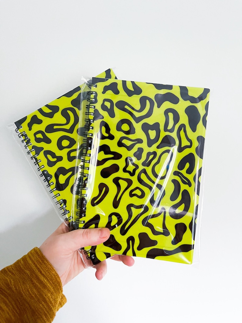 May include: Two spiral bound notebooks with black and yellow abstract patterns. The notebooks are wrapped in clear plastic.