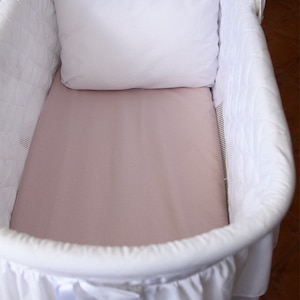 Puede incluir: Un moisés de mimbre blanco con una falda blanca con volantes y una sábana bajera rosa. Una almohada blanca está dentro del moisés.