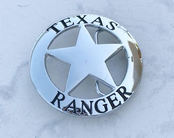 May include: A silver belt buckle with a black five-pointed star and the words "TEXAS RANGER" around the edge.