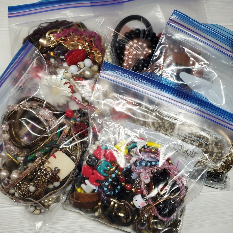May include: Four clear plastic bags filled with various colorful beads, charms, and jewelry. The bags are sealed with blue and white zipper closures.