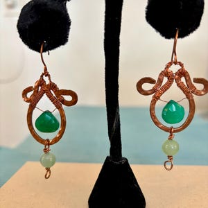 May include: A pair of copper wire earrings with green gemstone accents. The earrings feature a teardrop-shaped green stone, a smaller round green stone, and intricate copper wire designs.
