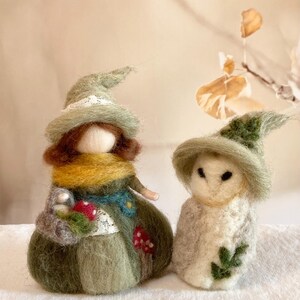 May include: Two felted figures: a witch-like doll with a green hat, scarf, and dress, holding a basket of mushrooms, and a smaller owl-like figure with a green hat and leaf accents. The figures are handmade and whimsical.