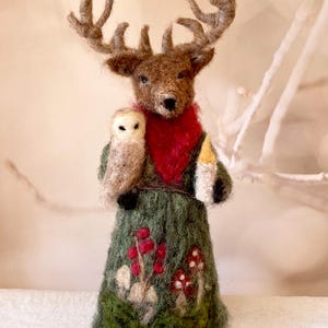 Puede incluir: Una figura artesanal de fieltro de una criatura parecida a un ciervo con grandes cuernos, una bufanda roja y una túnica verde adornada con bayas rojas y setas. Sostiene un pequeño búho y una vela.