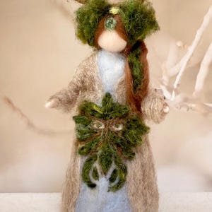 Puede incluir: Muñeca de fieltro hecha a mano que representa un espíritu del bosque. La muñeca presenta una máscara de hojas verdes, un abrigo marrón y una cabeza adornada con astas y follaje verde. El cuerpo de la muñeca es azul claro y blanco.