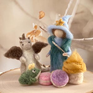 May include: A whimsical felted scene featuring a dragon, a wizard with a wand, a frog, a mushroom, and two macarons. The dragon has brown wings, and the wizard wears a blue hat. The scene is set on a wooden slice.