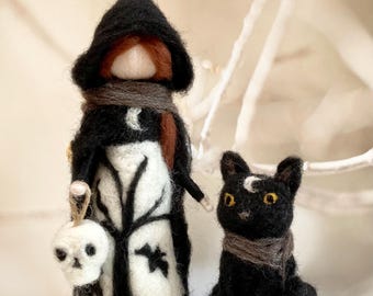 Può includere: Una bambola di strega in feltro con un mantello nero con cappuccio, che tiene una lanterna a forma di teschio, accanto a un gatto nero con un segno a mezzaluna. Entrambe le figure indossano sciarpe. L'abito della strega presenta un disegno ad albero e pipistrello.