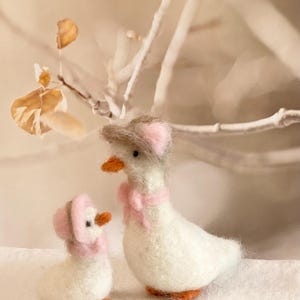 Puede incluir: Dos gansos de fieltro blanco con sombreros y bufandas rosas. El ganso más grande está de pie y el más pequeño está sentado.