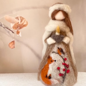 May include: A handcrafted felt doll with a winter theme. The doll wears a white coat and hat, holding a candle. The skirt features an orange fox, a red-berried branch, and a small bird. The doll has long brown hair.