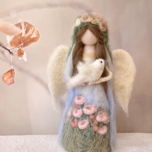 Puede incluir: Muñeca de ángel de fieltro hecha a mano con un vestido azul claro adornado con flores rosas y hierba verde. El ángel tiene alas blancas, una corona floral y sostiene una paloma blanca. Artículo decorativo.