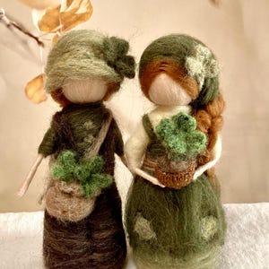 Könnte beinhalten: Zwei Filzpuppen in grüner Kleidung mit braunen Haaren und grünen Hüten. Die Puppen halten grüne Pflanzen in braunen Töpfen. Die Puppen stehen auf einer weißen Oberfläche.