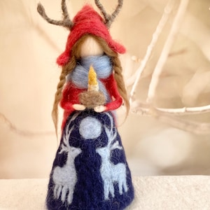 Könnte beinhalten: Handgefertigte Filzpuppe mit rotem Kapuzenmantel und braunen Zöpfen. Die Puppe hält eine Kerze und trägt einen blauen Schal. Der dunkelblaue Rock zeigt einen Mond und zwei hellblaue Rentiere.