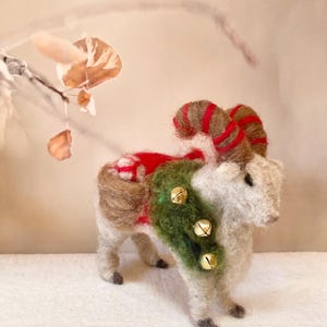 May include: A handmade felt ram figure, crafted from brown and white wool. The ram features red and brown striped horns, a green garland adorned with three gold bells, and a red saddle. A festive holiday decoration.