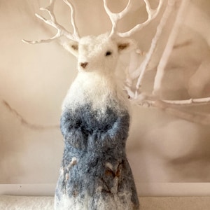 Op de afbeelding: Een naaldgevilt hertfiguurtje met een witte kop en groot wit gewei, overgaand in een textuurrijk lichaam met gemengde tinten blauw, grijs en witte wol. De onderkant van het figuurtje vertoont subtiele bruine patronen die op takken lijken.