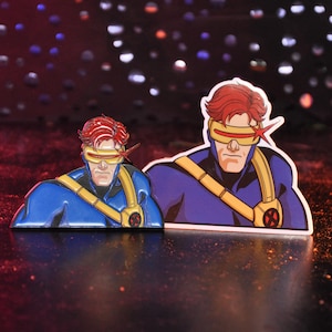 May include: A metal enamel pin and a matching vinyl sticker featuring a comic book character with red hair, a yellow visor, and a blue suit with a yellow strap and a circular red and black emblem on the chest, depicted in a heroic pose.