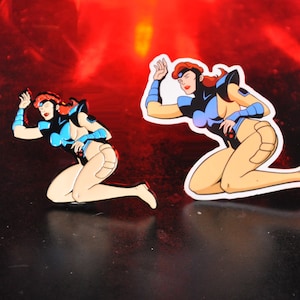 May include: Two enamel pins featuring a female superhero character in a blue and black costume. The character is kneeling with her arms raised. The pins are on a black surface with a red background.