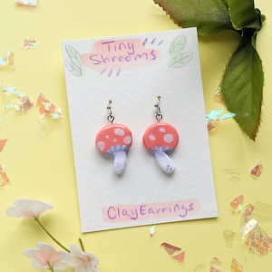 May include: A pair of clay earrings shaped like red and white mushrooms with purple stems. The earrings are on a white card with the text "Tiny Shrooms" and "Clay Earrings".