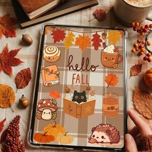 Puede incluir: Una tableta en una funda protectora transparente con una ilustración digital sobre un fondo de cuadros beige, hojas de otoño, un rollo de canela, una taza, un libro, una vela, calabazas, un erizo y un gato negro leyendo, con el texto hello FALL.