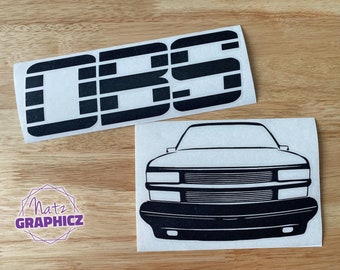 May include: Two black vinyl decals on a wooden surface. One decal features the letters "OBS" in a bold, blocky font. The second decal is a front view illustration of a vehicle. A purple and white logo for "Natz GRAPHICZ" is also visible.