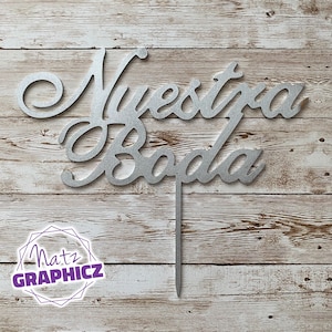 May include: Silver cake topper with the words "Nuestra Boda" in a cursive font. The topper is on a wooden background.