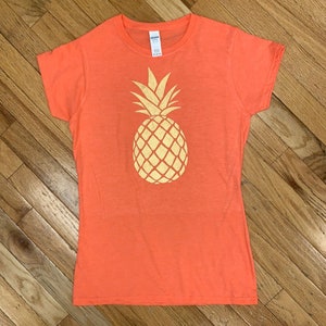 Peut inclure: Un t-shirt orange corail avec un motif d'ananas doré.
