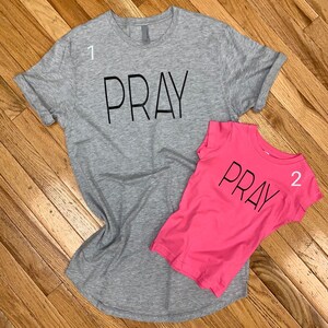 May include: Two t-shirts with the word "PRAY" printed on them. The larger shirt is grey and the smaller shirt is pink.