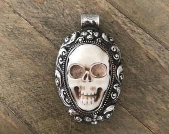 May include: Oval pendant featuring a skull design. The skull is white with brown eye sockets and a detailed jaw. The pendant is set in a silver-colored frame with ornate scrollwork and a bail for a chain.