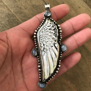 May include: A silver-toned pendant in the shape of a wing, featuring a mother-of-pearl inlay and three small, oval-shaped blue gemstones. The pendant has a detailed, textured design and a loop at the top for a chain. The wing design is bordered by small silver beads.