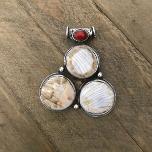 May include: A silver pendant featuring three round, white and brown shell pieces with a red stone at the centre.