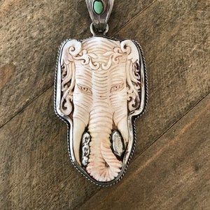 May include: A silver pendant featuring a carved white elephant head with a turquoise stone accent. The pendant has a detailed design and is framed with a silver rope border. "PENDANT-1" is written in the top right corner of the image.
