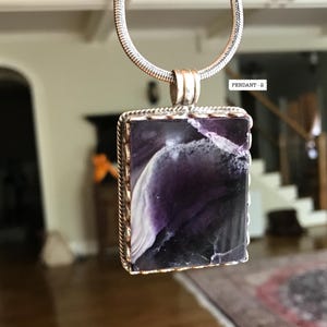 May include: A silver chain necklace with a rectangular pendant featuring a purple and white stone with a silver border. The pendant has a scalloped edge.