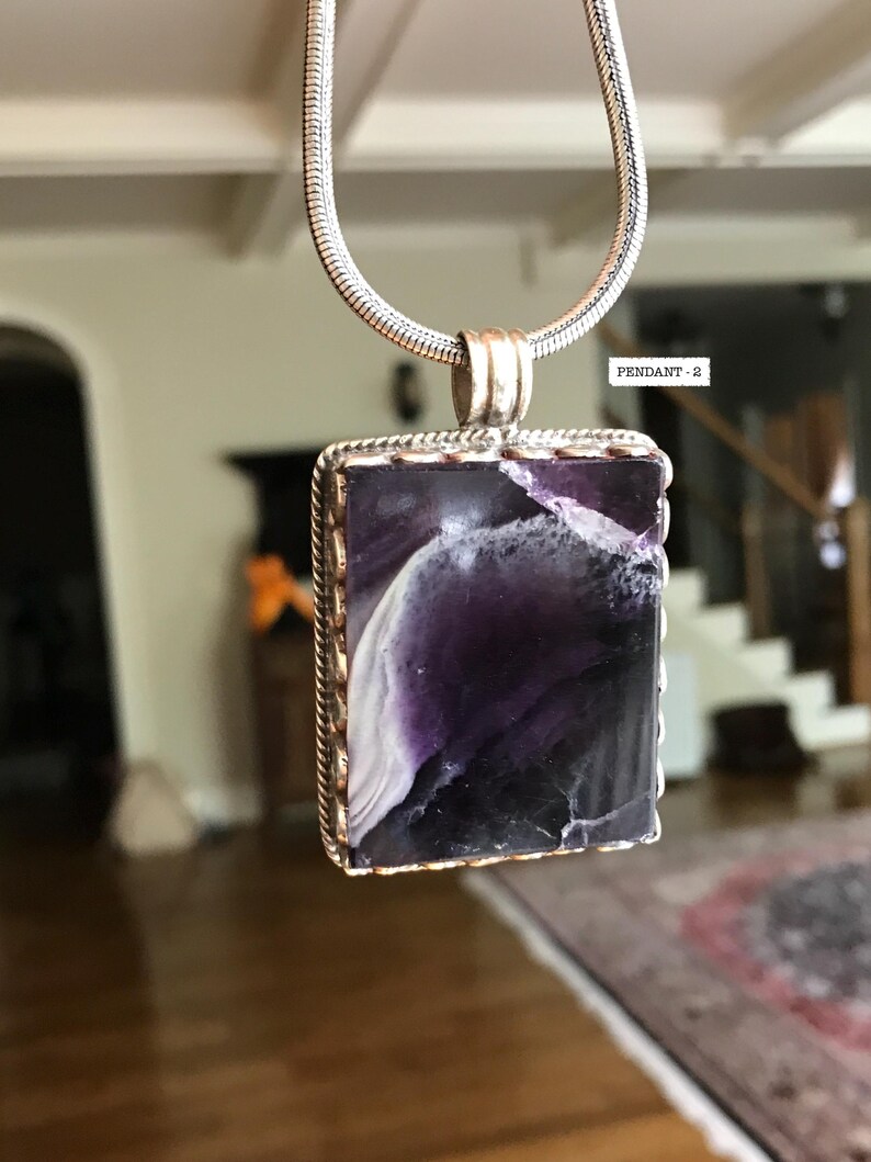 May include: A silver chain necklace with a rectangular pendant featuring a purple and white stone with a silver border. The pendant has a scalloped edge.